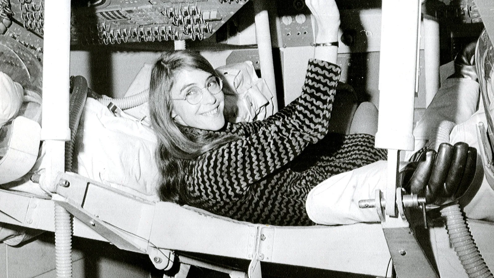Margaret Hamilton sorri deitada no assento do módulo de comando da Apollo, com o painel acima