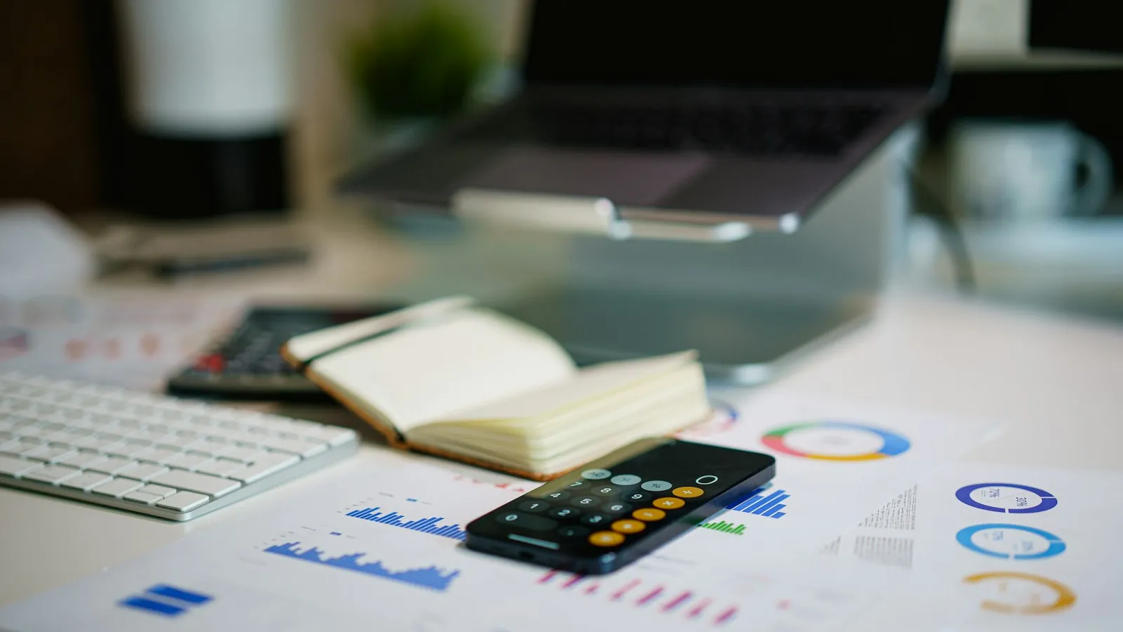 Mesa de escritório com celular aberto na calculadora, caderno, teclado e relatórios com gráficos financeiros