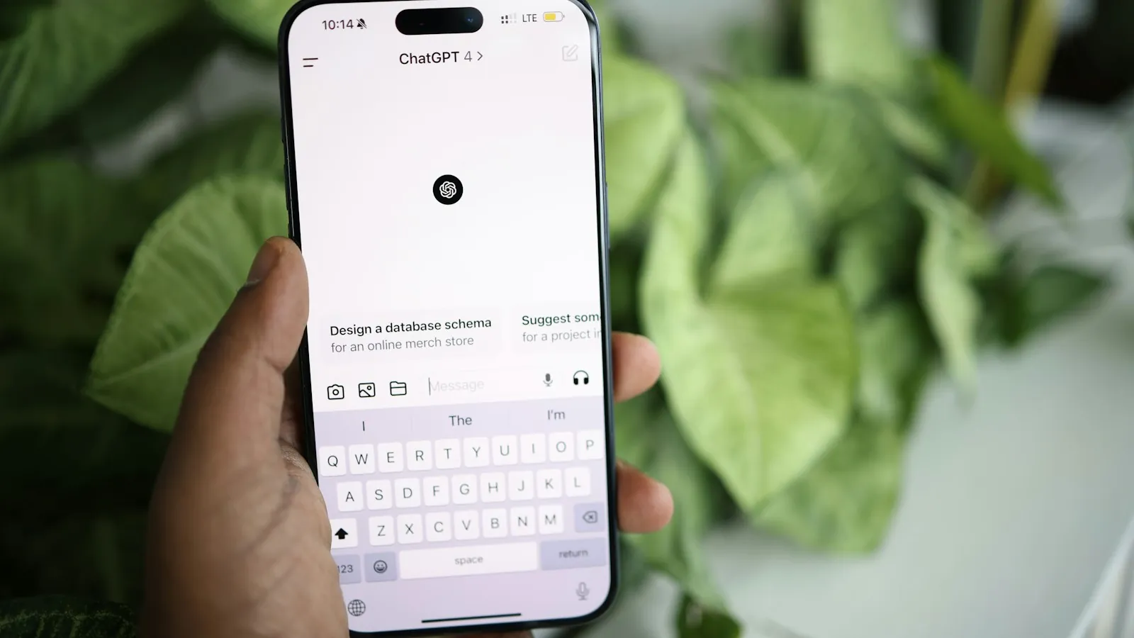 Mão segura celular com o aplicativo do ChatGPT aberto na tela de nova conversa, com folhagens ao fundo
