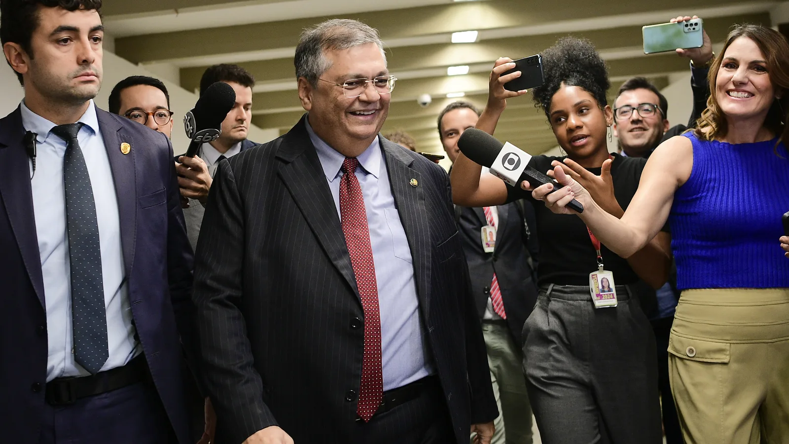 Flávio Dino sorri ao caminhar entre jornalistas e seguranças num corredor