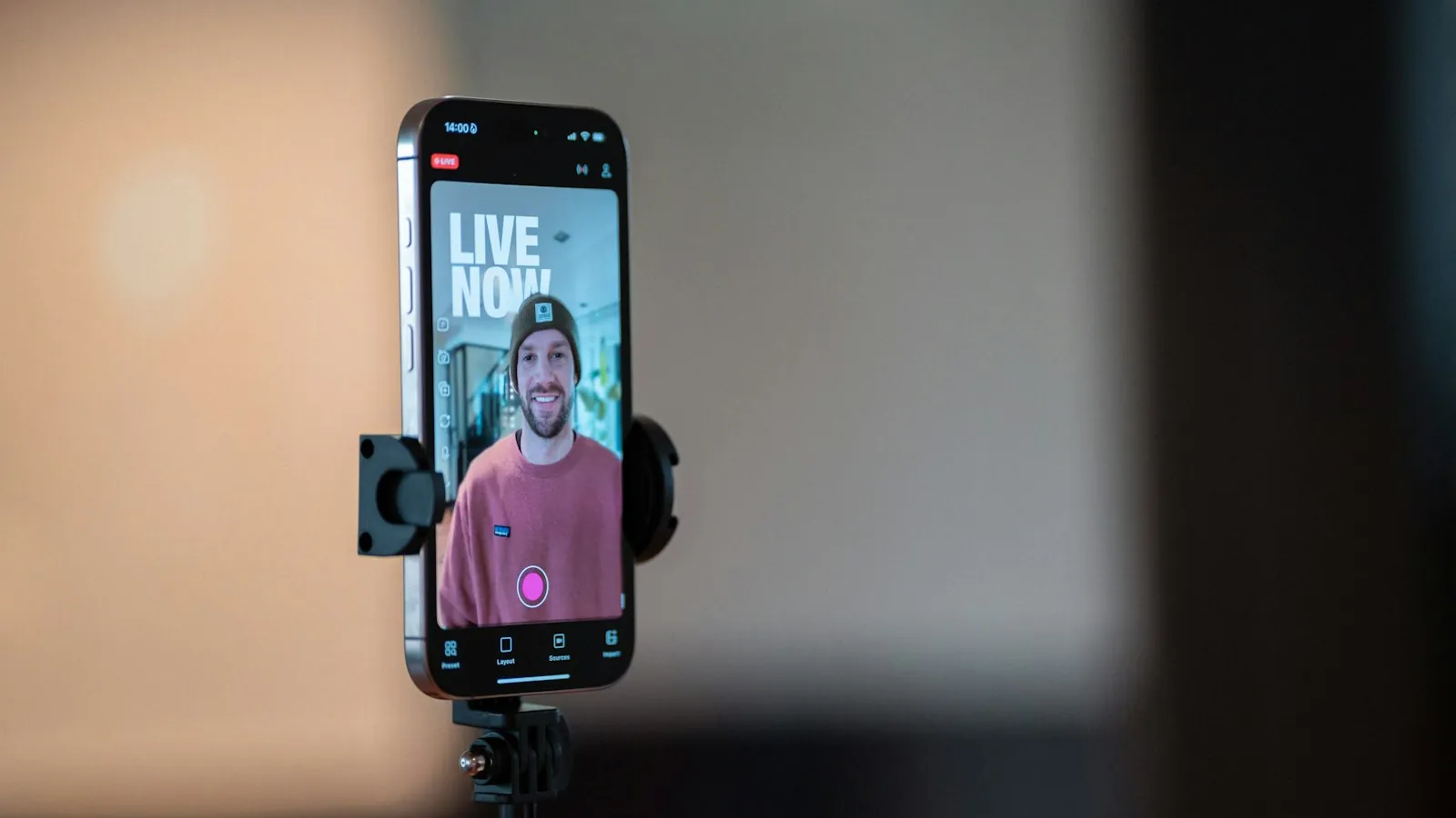 Celular preso a um tripé exibe transmissão ao vivo de um homem sorridente de gorro, com selo vermelho de live na tela