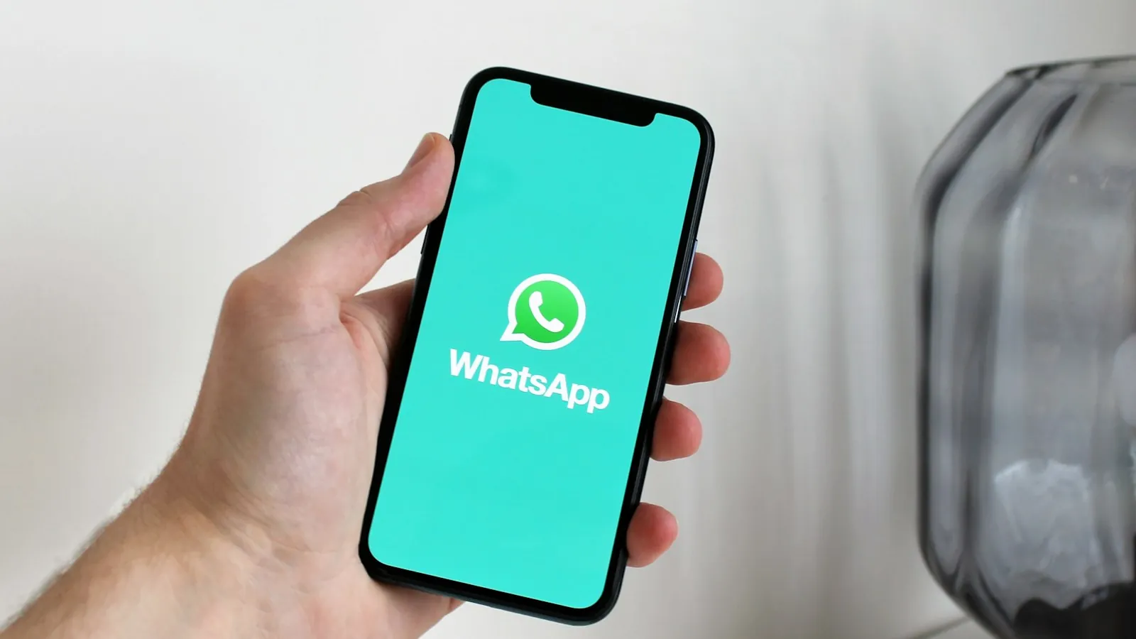 Mão segura celular com a tela de abertura do WhatsApp, com logo e nome do app sobre fundo verde-água